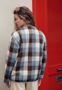 Florystyczna kurtka polarowa w kratę w kolorach niebieskim, białym i brązowym z zaokrąglonym dekoltem. Posiada luźny krój i ściągacze na mankietach. Tło jest czerwone.