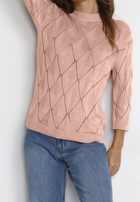 Rosa gestrickter Pullover mit Rautenmuster, 3/4-Ärmeln und geripptem Ausschnitt. Kombiniert mit blauen Jeans ist der strukturierte Stoff deutlich sichtbar.