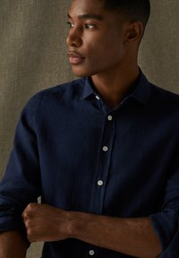 Námornícka košeľa na gombíky vyrobená z textúrovanej látky. Obsahuje klasický golier, biele gombíky a zvinutý rukáv, s jemnými vyšívanými detailmi.