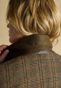 Pruun ruuduline jakk tekstureeritud villase kraega, millel on reljeefne sõna "JOULES" ja siledast nahast aktsent kaela ümber.