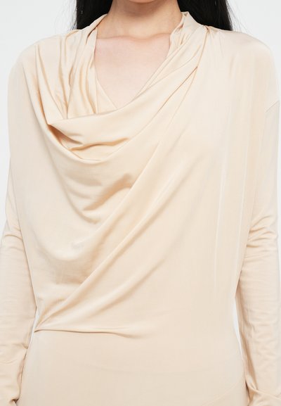Beige drape-top, jossa on pitkät hihat, pehmeä ja virtaava kangas sekä kaula-aukko, joka luo lempeitä poimuja rinnan ympärille.
