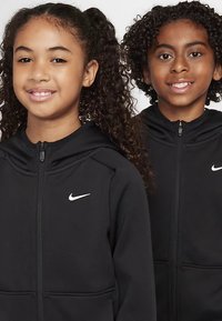 Doi copii purtând hanorace negre cu fermoar, având un mic logo alb Nike în partea stângă a pieptului. Amândoi au păr creț și zâmbesc.