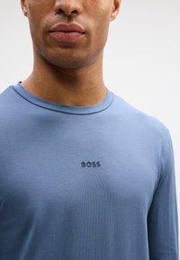 Hosszú ujjú kék póló sima anyagból, kerek nyakkivágással és egy kis fekete "BOSS" logóval a mellkasán.