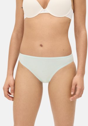 Mujer con sujetador blanco y ropa interior azul pastel claro, mostrada desde la mitad del torso hasta la parte superior de los muslos contra un fondo liso.