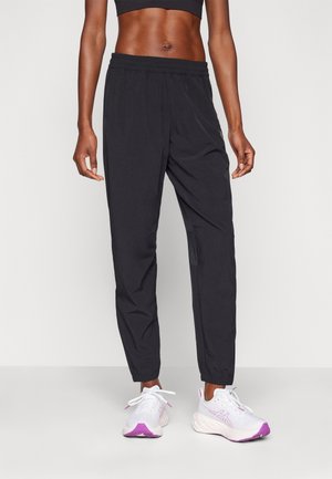 Persona atlética con pantalones jogger negros y zapatillas de running blancas con detalles rosas, de pie frente a un fondo claro y liso.