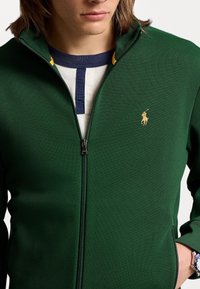 Grön zip-up jacka av texturerat tyg, med en guldfärgad logotyp på vänster bröst och en krage i marinblått och gult.