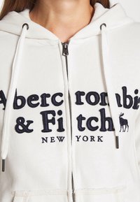 Bílá mikina na zip s kapucí, s navy vyšívaným textem "Abercrombie & Fitch" a grafikou losa; obsahuje stahovací kapuci a kovový zip.