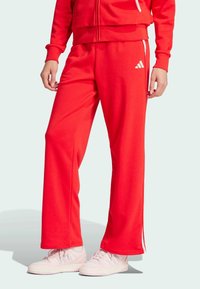 Crvene trenirke napravljene od mekanog materijala, s opuštenim krojem, bijelim bočnim prugama i Adidas logom na lijevoj nozi.