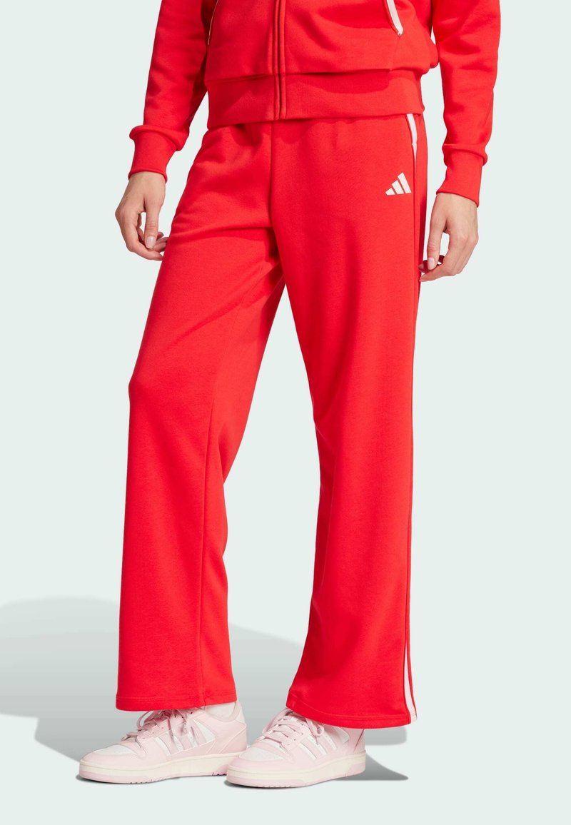 Crvene trenirke napravljene od mekanog materijala, s opuštenim krojem, bijelim bočnim prugama i Adidas logom na lijevoj nozi.