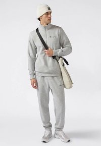 Szary polarowy pulower z półzamkiem i haftowanym logo, zestawiony z dopasowanymi dresami. Akcesoria to biała dziana czapka i beżowa torba.