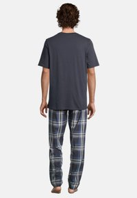 Man med lockigt hår står barfota med ryggen mot kameran, iklädd en mörkgrå kortärmad skjorta och blå rutig pyjamasbyxor.
