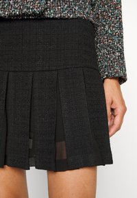 Gros plan d'une jupe plissée noire texturée associée à une veste en tweed multicolore, montrant une main reposant à côté de la jupe.