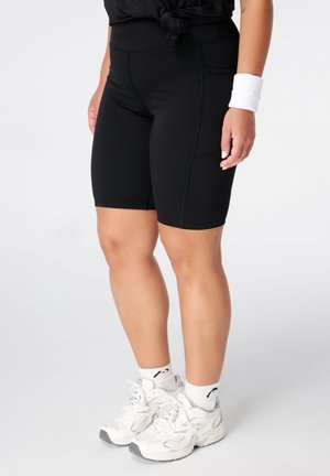 Personne portant un short de cyclisme noir jusqu'aux genoux, des chaussures de sport blanches, des chaussettes blanches et un bracelet blanc sur un fond uni.