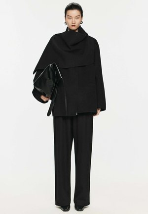 Black wool coat with a draped design, paired with wide-leg black trousers and a structured, glossy black bag. Smooth, textured surfaces.