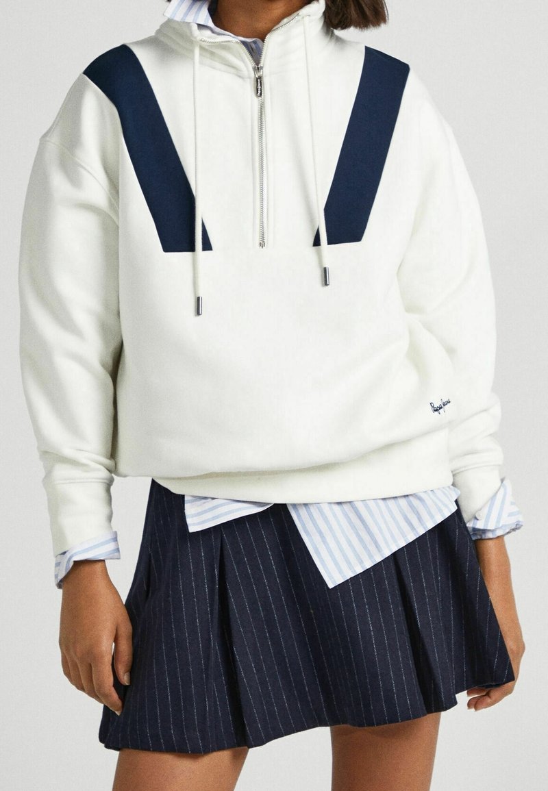 Sweat à capuche blanc à demi-fermeture éclair avec détails en chevrons bleu marine et cordons de serrage, porté sur une chemise rayée bleue, associé à une jupe plissée à fines rayures bleu marine.