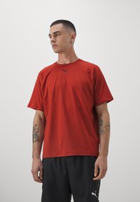 Vyras su trumpais tamsiais plaukais ir tatuiruotėmis, dėvintis raudoną Puma marškinėlį ir juodas Puma šortus, žiūrintis į kairę priešais paprastą foną.