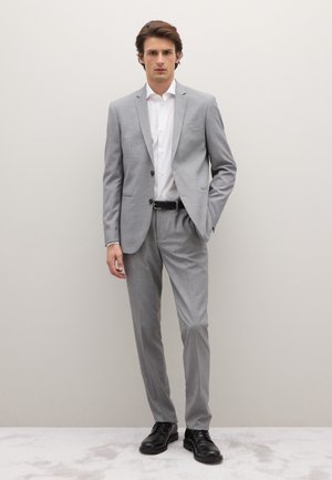 Completo grigio chiaro su misura con giacca e pantaloni aderenti. Camicia bianca elegante, cintura nera e scarpe eleganti nere. Tessuto liscio.