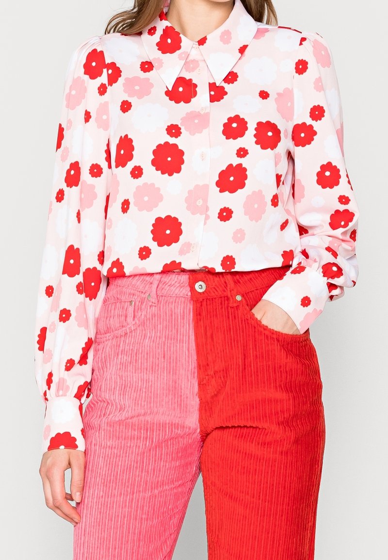 Blommig blus med röda och rosa blommor på en ljus bakgrund, matchad med corduroybyxor som är rosa på ena sidan och röda på den andra.