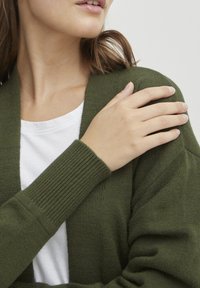 Cardigan en laine verte tricoté avec des poignets côtelés et une ouverture sur le devant. Porté par-dessus un t-shirt blanc, mettant en valeur sa texture douce et sa coupe décontractée.
