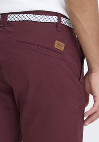 Pantalons bordeaux avec une ceinture à motif blanc et bleu, dotés d'un patch en cuir sur la poche arrière, mettant en valeur une texture de tissu lisse.