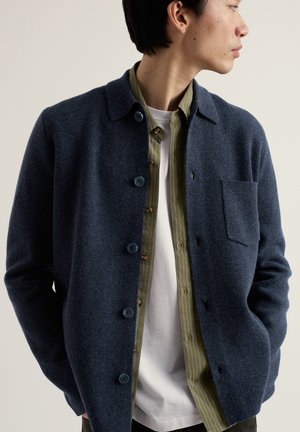 Jeune homme portant une veste en laine bleue marine boutonnée sur une chemise rayée verte et un t-shirt blanc, faisant face à droite avec les mains dans les poches.