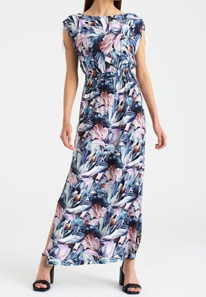 Kvinde iført en ærmeløs, gulvlang kjole med blomsterprint, rynket talje og sideslids, parret med sorte sandaler med hæl.