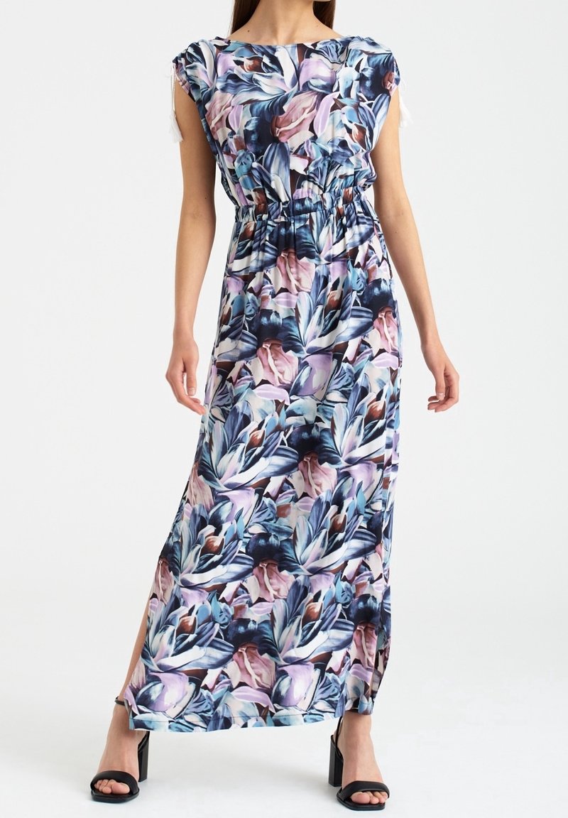 Femme portant une robe longue à imprimé fleuri, sans manches, à taille froncée et fente sur le côté, associée à des sandales noires à talons.