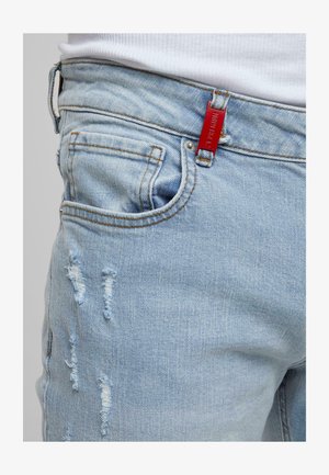 Jeans in denim blu chiaro con effetto usurato e toppe visibili, tasca frontale, passante per la cintura e etichetta rossa del marchio in vita, indossati con una camicia bianca.