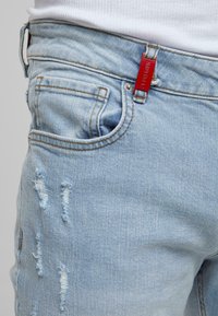 Light blue distressed denim jeans with frayed patches, orange stitching, five pockets, and a prominent red brand label on the waistband.