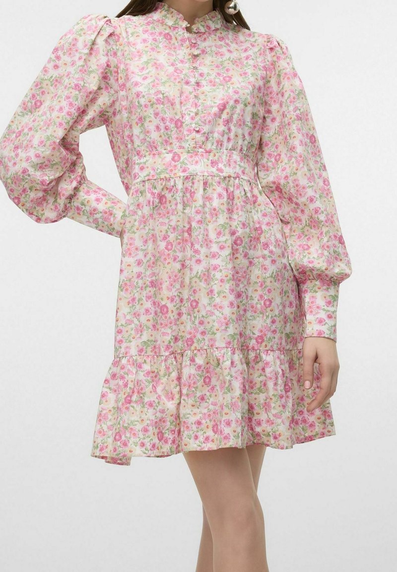 Kvinna i en långärmad rosa blommig klänning med puffiga axlar och en volangkant, poserande med en hand på höften.