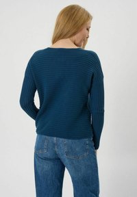 Dunkeltürkiser gerippter Pullover mit langen Ärmeln, weitem Ausschnitt und geradem Saum, kombiniert mit hellblauen Jeans. Texturierte Strickdetails sichtbar.