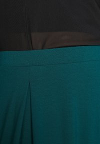 Svart meshmateriale i toppen, som övergår till en solid mörk teal-nedre del med en veckdetalj och en slät textur.