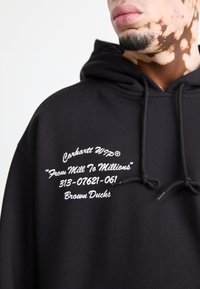 Felpa nera con cappuccio realizzata in tessuto spesso, caratterizzata da testo ricamato bianco: "Carhartt WIP® Da Mulino a Milioni" e "Anatre Marroni". Include cordini.