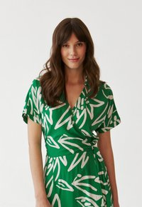 Grünes Wickelkleid aus leichtem Stoff mit weißen abstrakten Blumenmustern, kurzen Ärmeln und einem Bindeband in der Taille.