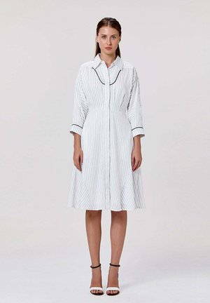 Femme portant une robe blanche mi-longue à fines rayures verticales, bordure noire au col et aux poignets, et escarpins noirs et blancs à bride cheville.