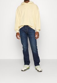 Sudadera con capucha amarilla, vaqueros de mezclilla azules y zapatillas blancas con detalles en verde. Conjunto casual con un corte relajado y detalles mínimos.