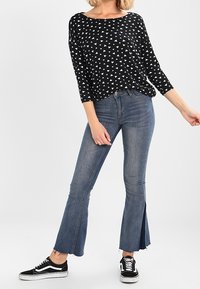 Chemise noire à motif triangulaire blanc, manches trois-quarts, associée à un jean bleu évasé avec un ourlet brut et des fentes latérales. Baskets noires.