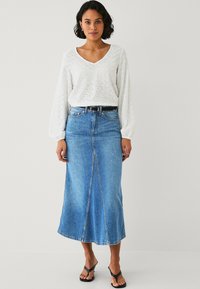 Blusa bianca strutturata con maniche lunghe, abbinata a una gonna di jeans blu a metà lunghezza con uno spacco frontale, accentuata da una cintura nera e sandali.