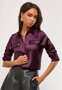 Satin, dark purple button-down shirt with a pointed collar, two chest pockets, and rolled sleeves, paired with a black leather skirt.