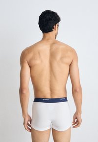 Homme aux cheveux courts et foncés, debout de dos, portant un boxer blanc avec une ceinture foncée portant l'inscription « GANT », sur un fond uni.