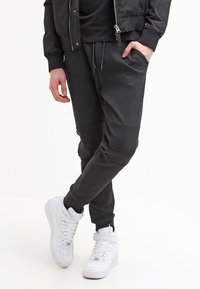 Man i svarta joggarbyxor och en svart jacka med en hand i fickan, tillsammans med vita högstövlar, står mot en vit bakgrund.