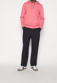 Homme portant un sweat à capuche rose, un pantalon noir et des baskets blanches avec des bandes noires, debout les mains dans les poches devant un fond uni.