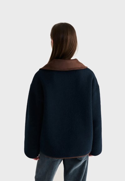 Femme vue de dos portant un manteau bleu marine foncé avec un col marron et un pantalon grisâtre sur un fond clair uni.