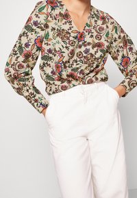 Beige Bluse mit Blumenmuster, V-Ausschnitt und geknöpften Ärmeln, kombiniert mit weißen, hoch taillierten Hosen. Besitzt lebendige Farben und Details.