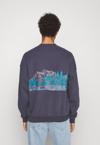 Granatowa bluza z grafiką gór i drzew w odcieniach teal i różu na plecach, o wygodnym, luźnym kroju.