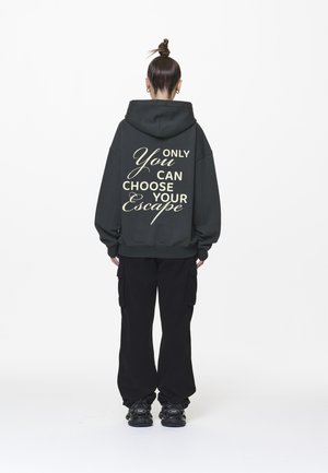Personne avec un chignon, portant un sweat à capuche foncé avec le texte "Only You Can Choose Your Escape" dans le dos, un pantalon cargo noir et des baskets noires.