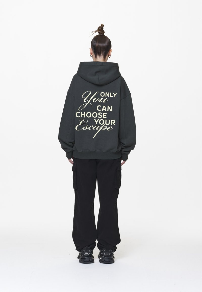 Personne avec un chignon, portant un sweat à capuche foncé avec le texte "Only You Can Choose Your Escape" dans le dos, un pantalon cargo noir et des baskets noires.
