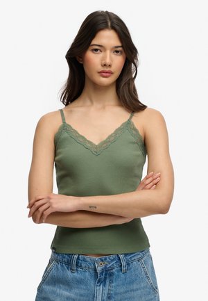 Kobieta o długich ciemnych włosach, ubrana w zielony top z koronkową lamówką i niebieskie jeansy, ze skrzyżowanymi ramionami, neutralny wyraz twarzy.