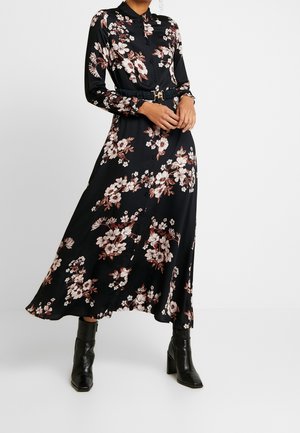 Femme portant une robe chemise à manches longues noire à fleurs, ceinturée, avec des bottines à talons noires, se tenant devant un fond uni.