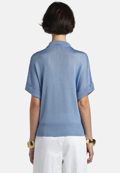 Femeie cu păr scurt și întunecat, purtând un top tricotat cu mâneci scurte de culoare albastru deschis și pantaloni albi, văzută de spate pe un fundal simplu.